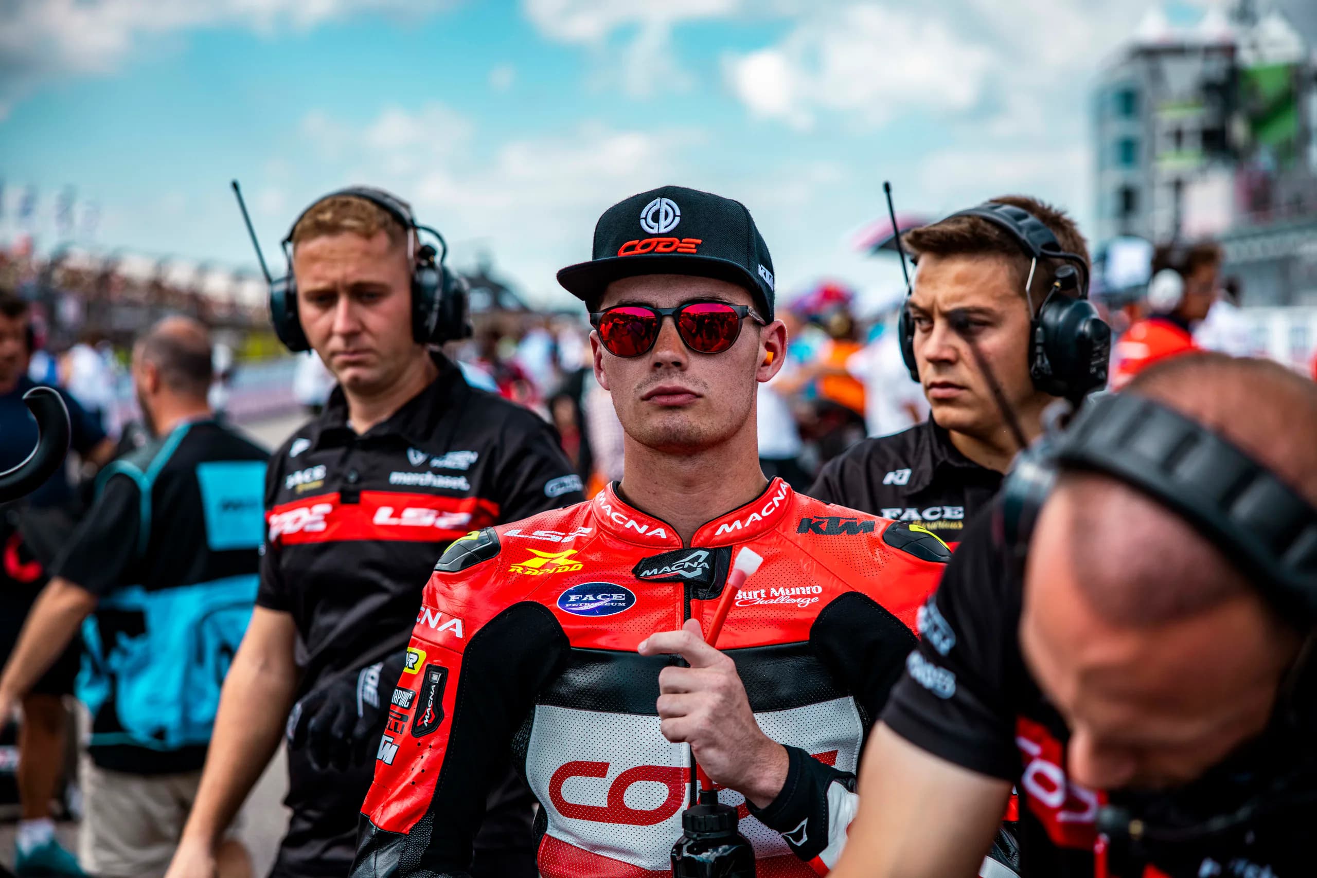 Cormac Buchanan in CODE team leathers and red sunglasses on the Moto3 grid, surrounded by crew in headsets