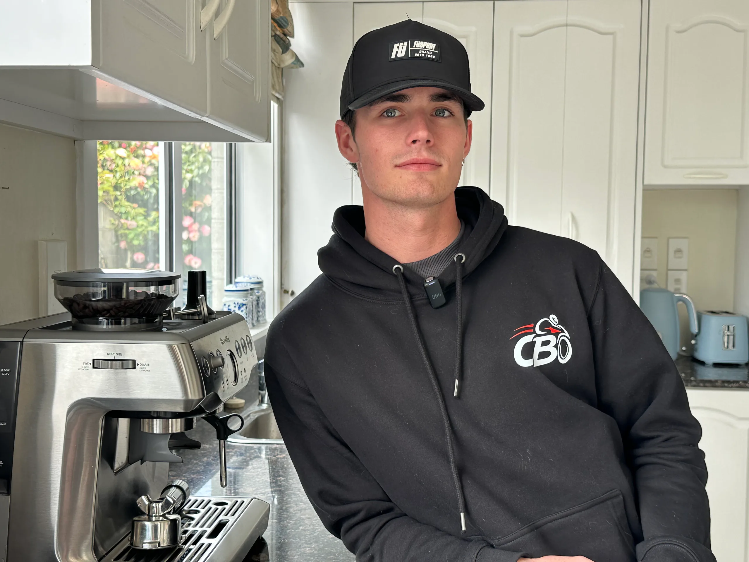 Cormac Buchanan in a black cap and hoodie standing beside an espresso machine in a kitchen