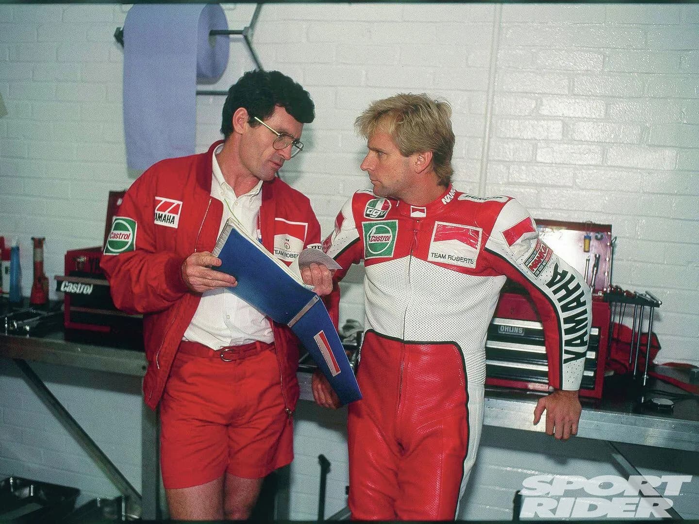 Mike Sinclair and Wayne Rainey in red Yamaha team gear looking over a folder in a garage